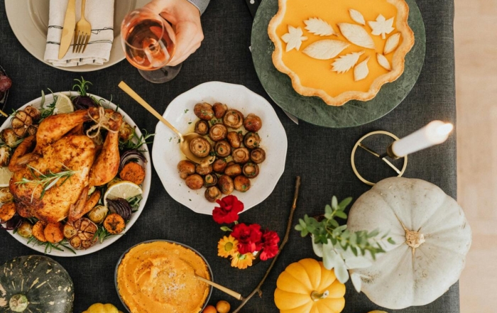 A table full of Thanksgiving dishes, a plate of whole turkey, a plate of mushrooms, mashed butternut squash, pumpkin pie, a glass of red wine, a lit up white candle, and pumpkins and flowers surrounding the table.