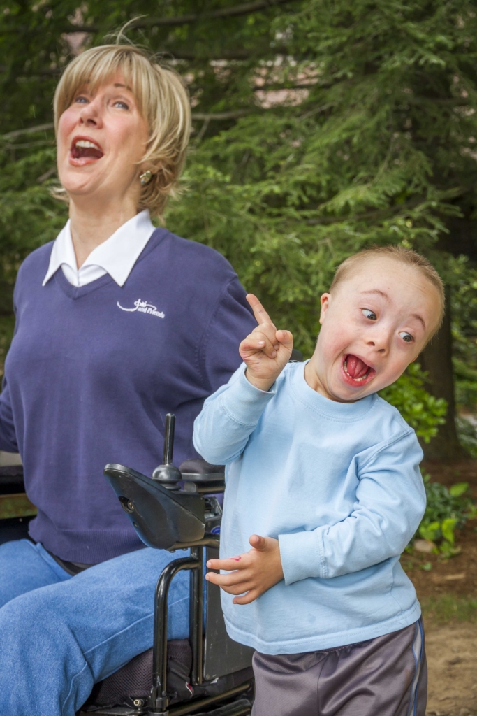 Joni Eareckson Tada, on the left, sits in her wheelchair with a joyful expression. Josiah stands next to her, pointing towards her with a beaming smile. The background features lush green trees, slightly blurred.