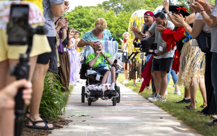 Buddy, sitting in his wheelchair, is being gently pushed by Sheila along a pathway at a Family Retreat. He holds his head as they move forward, surrounded by volunteers on both sides of the pathway who are greeting them with warm smiles and waves. The scene captures a moment of care and community support in a welcoming, outdoor setting.