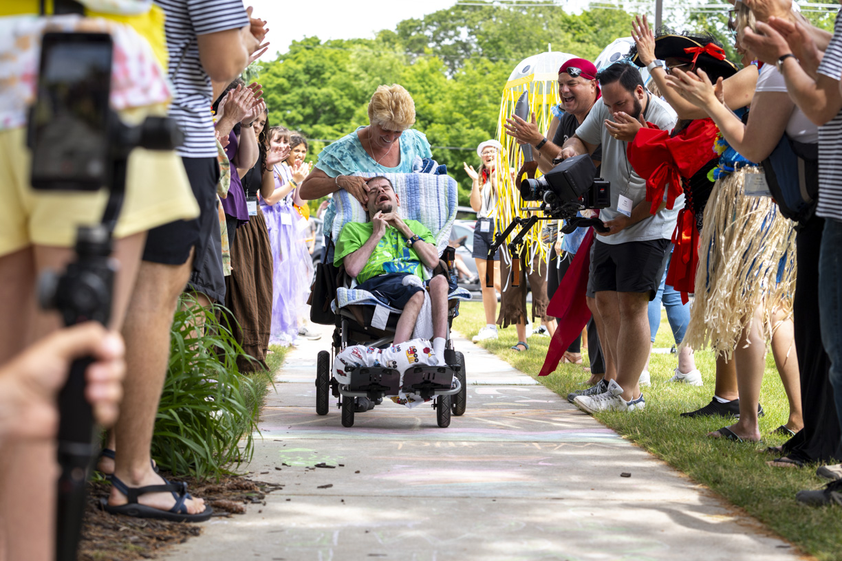 Buddy, sitting in his wheelchair, is being gently pushed by Sheila along a pathway at a Family Retreat. He holds his head as they move forward, surrounded by volunteers on both sides of the pathway who are greeting them with warm smiles and waves. The scene captures a moment of care and community support in a welcoming, outdoor setting.