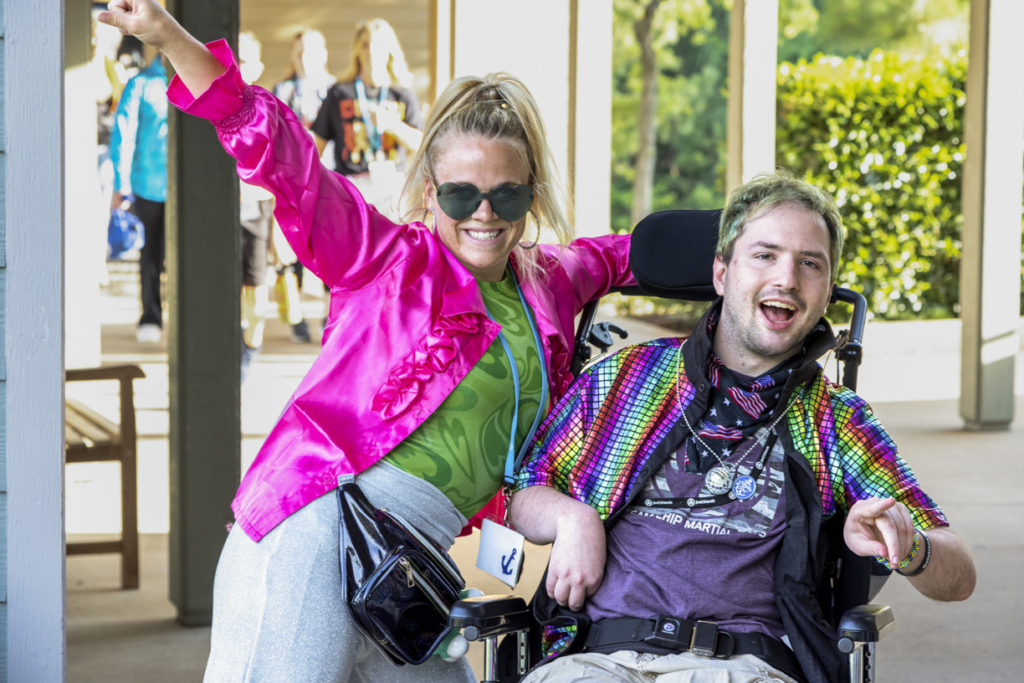Juddy is sitting in his wheelchair, wearing a colorful disco outfit, next to his buddy volunteer before Disco Night. They both look excited and full of energy!