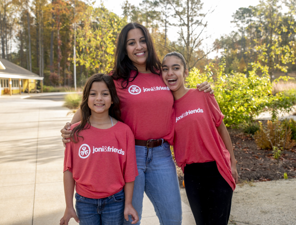 A picture of the Rodriguez family: Amelia on the left, Lorna in the middle, and Natalia on the right. They are all wearing red shirts with the Joni & Friends logo, standing and smiling for the camera with tall trees in the background.