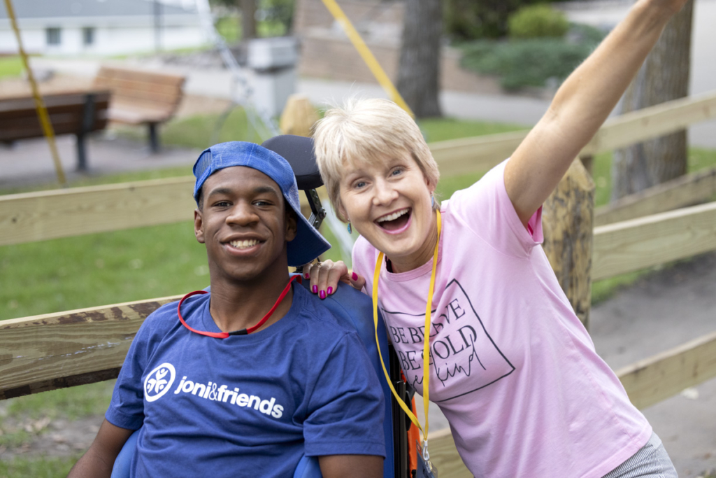 Josiah, wearing a blue t-shirt with the Joni & Friends logo next to his buddy at Family Retreat. They are smiling joyfully, surrounded by nature, and looking happy as they pose for the camera.