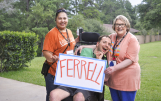 Juddy is in his wheelchair, positioned between Lanette and his caregiver, who are both holding a white paper with the word "Ferrell" written on it. They all look so excited.