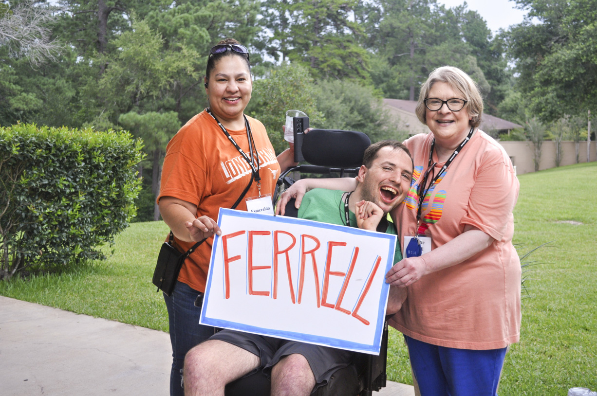 Juddy is in his wheelchair, positioned between Lanette and his caregiver, who are both holding a white paper with the word "Ferrell" written on it. They all look so excited.