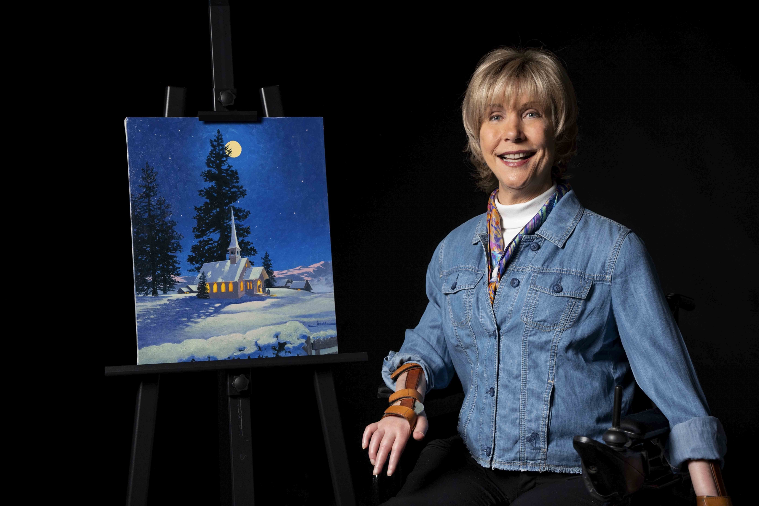 Joni Eareckson Tada wearing a button-up cardigan, a white turtleneck, and a colorful scarf, smiling for the camera next to her "Christmas Eve Night" artwork. She is set against a black background.