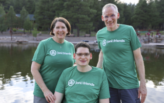 Traci on the left, Bryden in the middle sitting in his wheelchair, and Barry on the right, all wearing green t-shirts with the Joni & Friends logo. Behind them, there's a body of water and lush green trees.