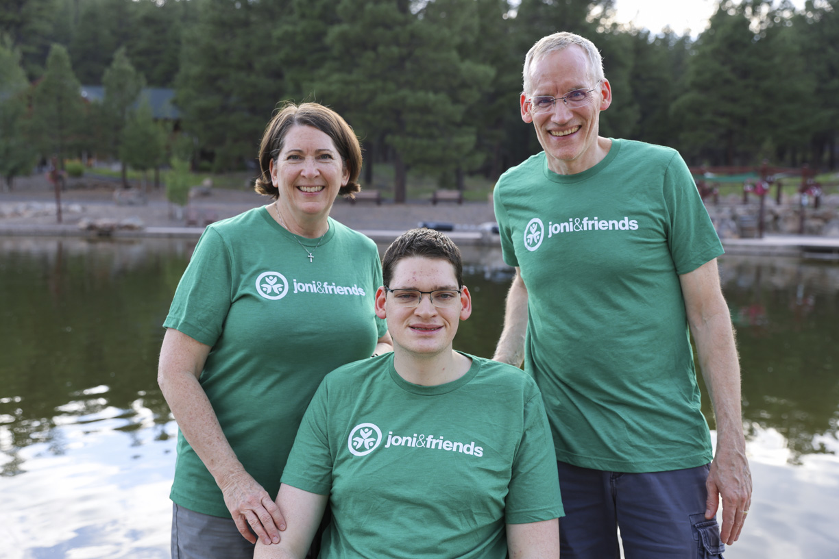 Traci on the left, Bryden in the middle sitting in his wheelchair, and Barry on the right, all wearing green t-shirts with the Joni & Friends logo. Behind them, there's a body of water and lush green trees.