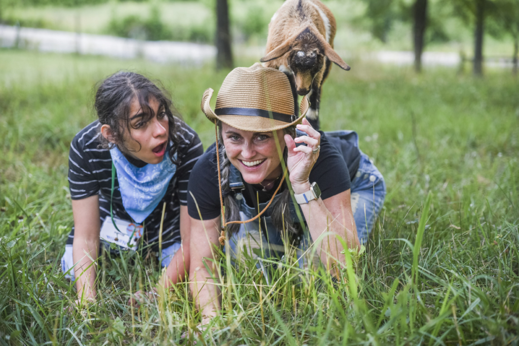 Natalia and her buddy smiling and kneeling on the grass during Family Retreat, with a goat standing behind them. 