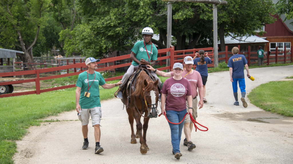 Josiah, wearing a green t-shirt with the Joni & Friends logo and a helmet, is happily riding a horse. He is surrounded by three Joni & Friends volunteers, who are assisting him with the ride, all smiling and engaged in the moment. 