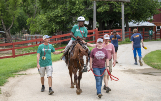 Josiah, wearing a green t-shirt with the Joni & Friends logo and a helmet, is happily riding a horse. He is surrounded by three Joni & Friends volunteers, who are assisting him with the ride, all smiling and engaged in the moment