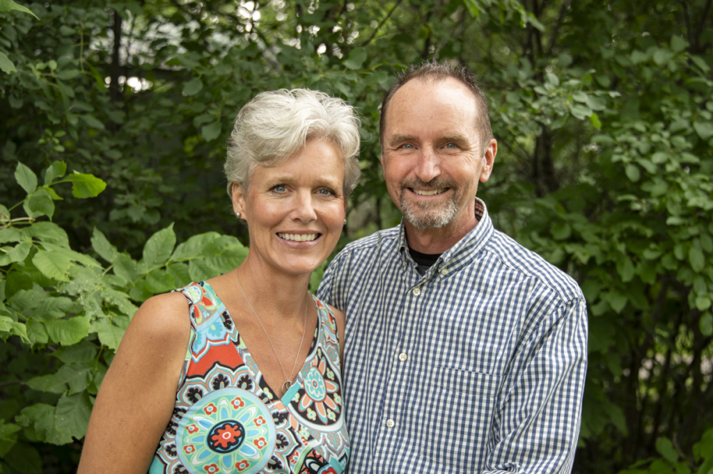 Christina and Scott standing next to each other, smiling and looking joyful as they pose for the camera. They are surrounded by a backdrop of lush trees, adding to the cheerful atmosphere.