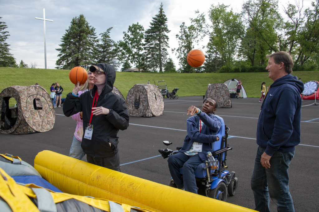 Josiah, in his wheelchair and next to his buddy, is focused as he throws a basketball. Nearby, another attendee, with their buddy, is also throwing a basketball, creating a moment of concentration and teamwork.