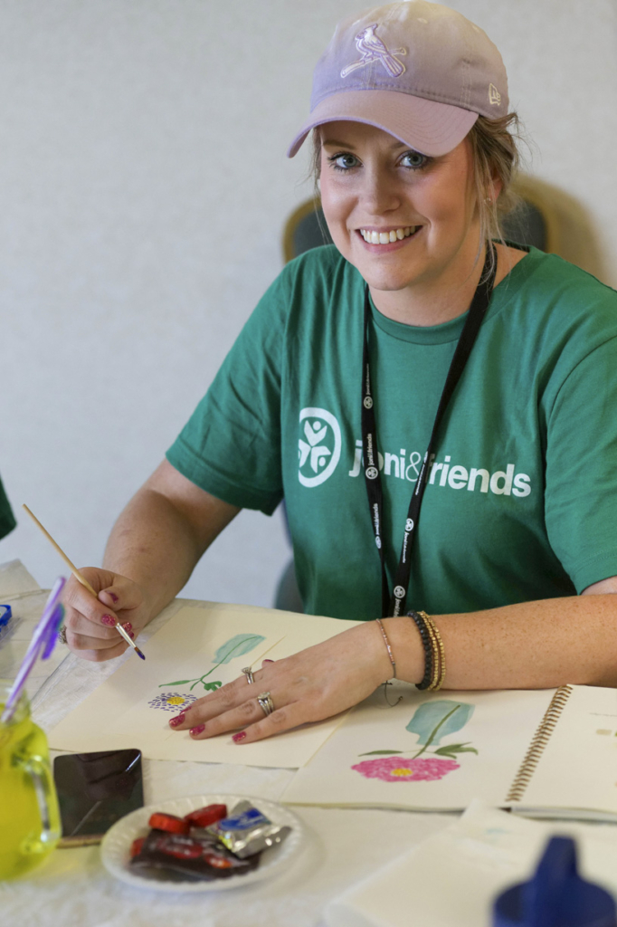 Brittany is smiling for the camera while holding a paintbrush in one hand. On the table in front of her are her beautiful paintings of vibrant flowers, showcasing her artistic talent.