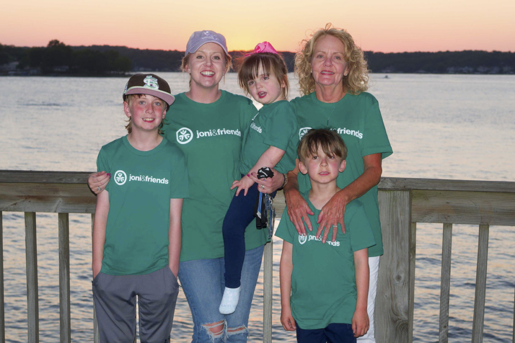 Brittany is being carried by her mom, surrounded by her brothers, Luke and Kolten, and her grandma, Maria. All are wearing matching green t-shirts with the Joni & Friends logo. The group is smiling and posing happily for the camera against a beautiful backdrop of a wooden deck railing and tranquil water during sunset, radiating joy and warmth.