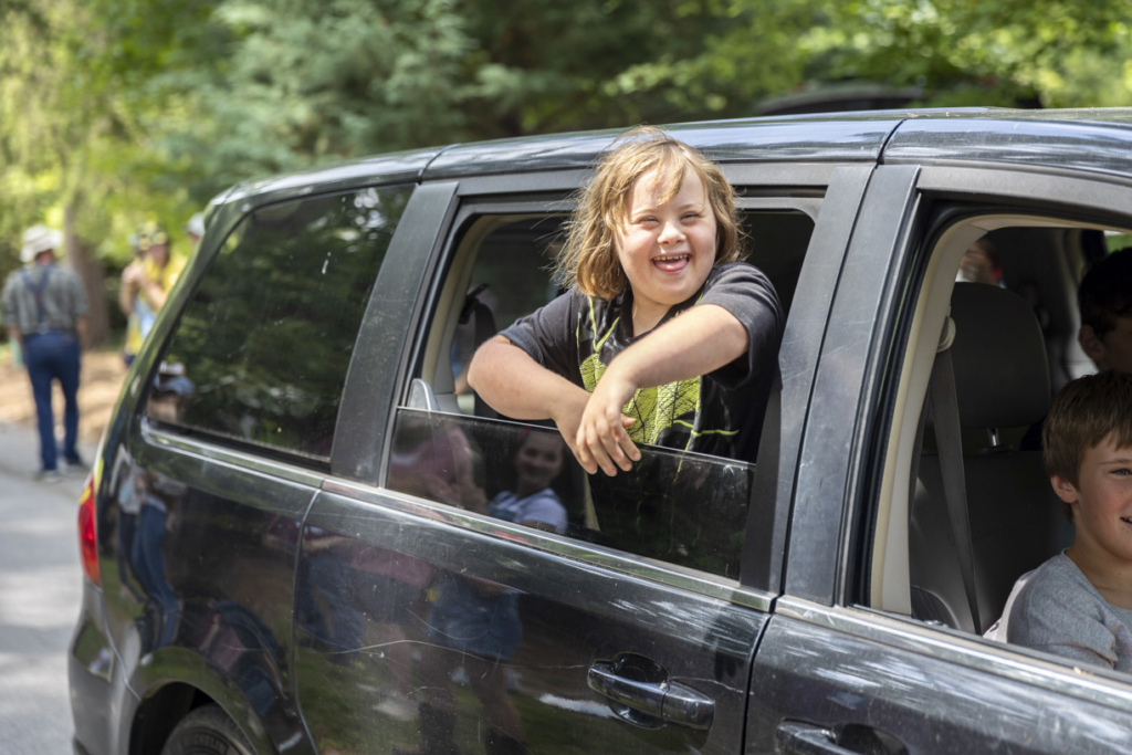 Ariella is excitedly leaning out of the car window, with her head and arms fully outside, wearing a big grin of happiness and excitement.