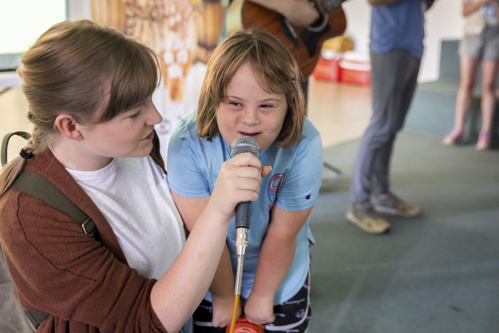 Ariella is singing into a microphone while a woman holds it for her. She looks very happy.