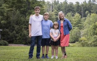 Anakin, Asher, Ariella, and Traci are standing together, posing for the camera on the grass, with tall, lush green trees in the background.