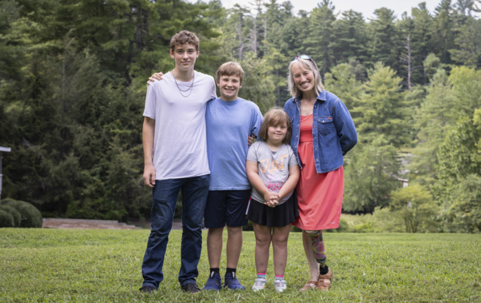 Anakin, Asher, Ariella, and Traci are standing together, posing for the camera on the grass, with tall, lush green trees in the background.