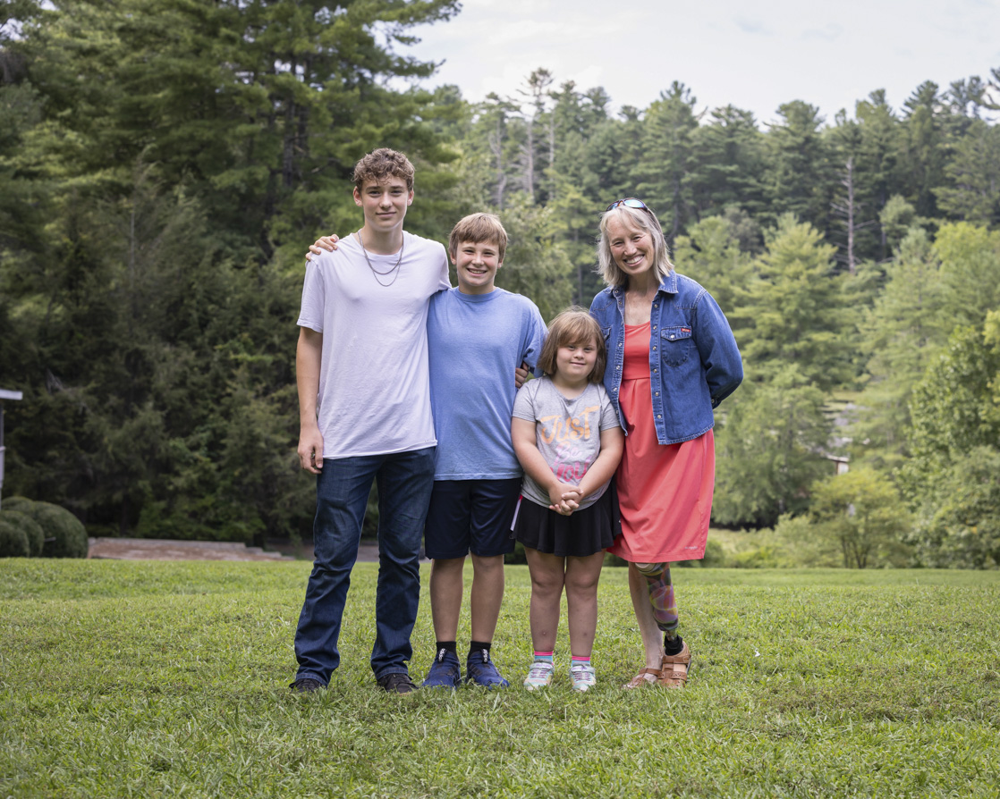 Anakin, Asher, Ariella, and Traci are standing together, posing for the camera on the grass, with tall, lush green trees in the background.