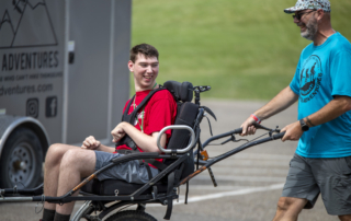 Gabe is sitting in a hiking wheelchair, looking excited as he is being pushed by a man wearing a blue t-shirt with a Luke5Adventures logo on it.