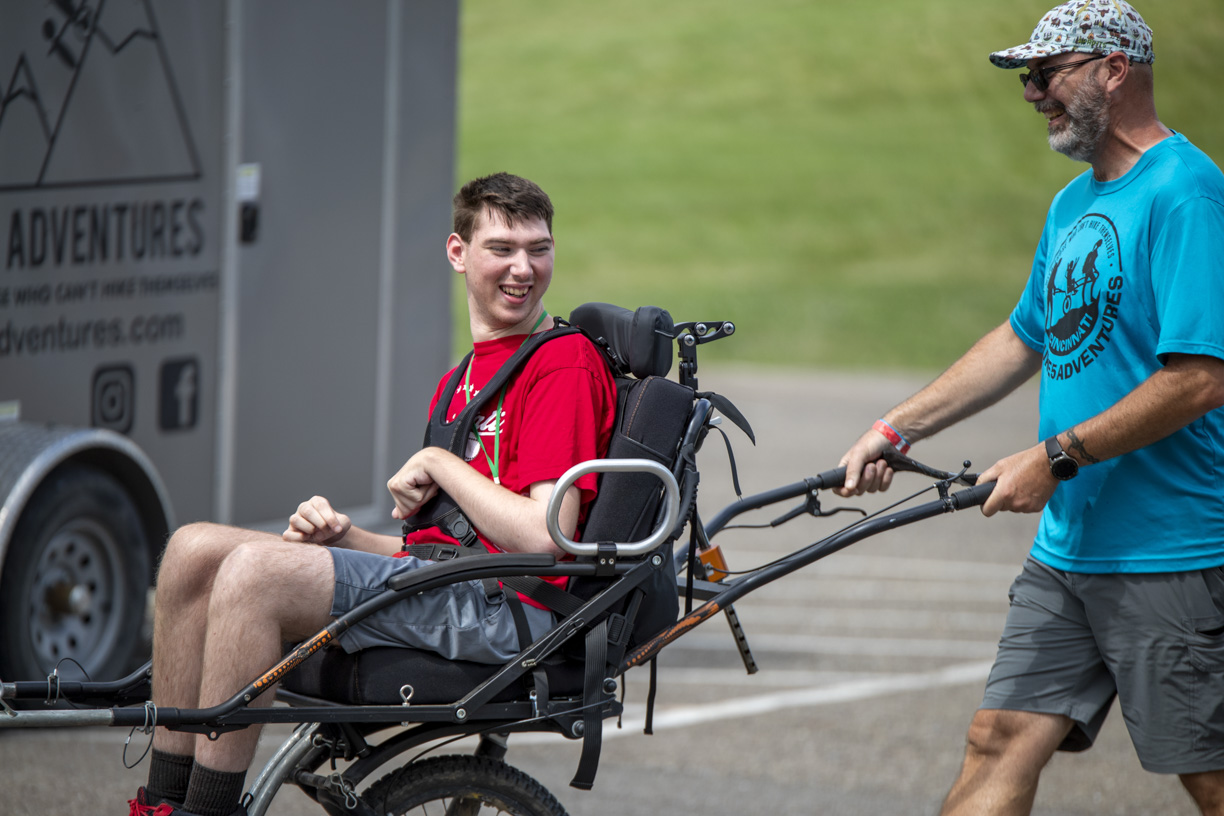 Gabe is sitting in a hiking wheelchair, looking excited as he is being pushed by a man wearing a blue t-shirt with a Luke5Adventures logo on it.