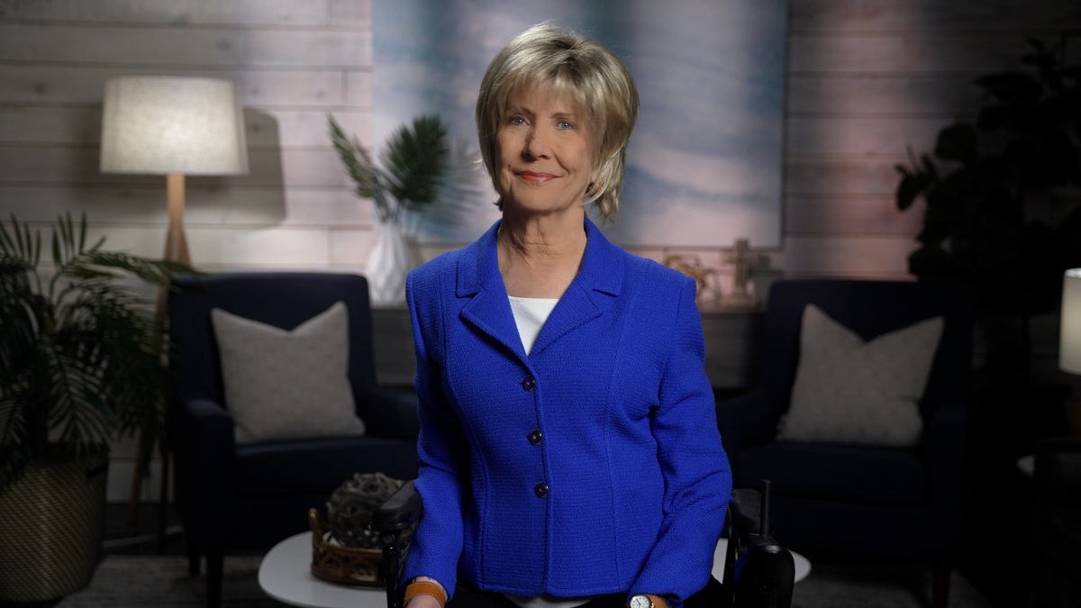 Joni Eareckson Tada is smiling for the camera, wearing a royal blue jacket over a white top. She is positioned in front of a softly blurred background that features a cozy living room setup.