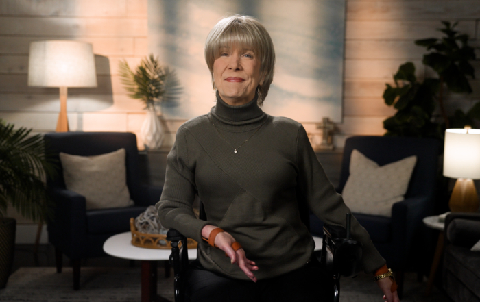 Joni wearing a turtleneck and a heart necklace is smiling for the camera. The background features a cozy living room setup.