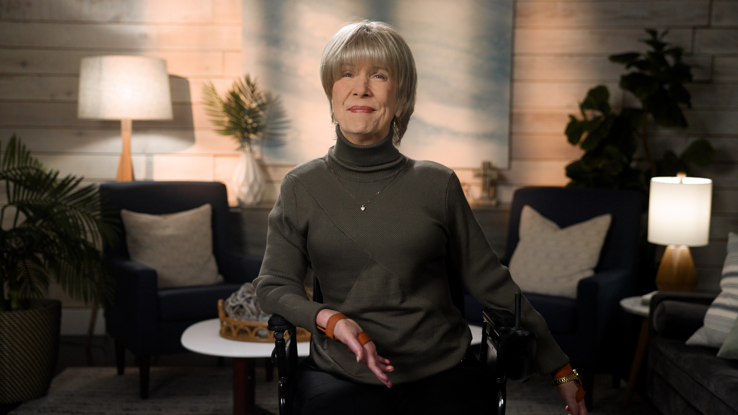 Joni wearing a turtleneck and a heart necklace is smiling for the camera. The background features a cozy living room setup.