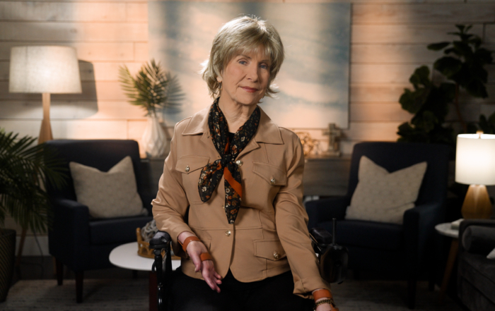 Joni Eareckson Tada smiling for the camera while wearing a brown safari jacket, a black scarf with floral details, in a cozy living room.