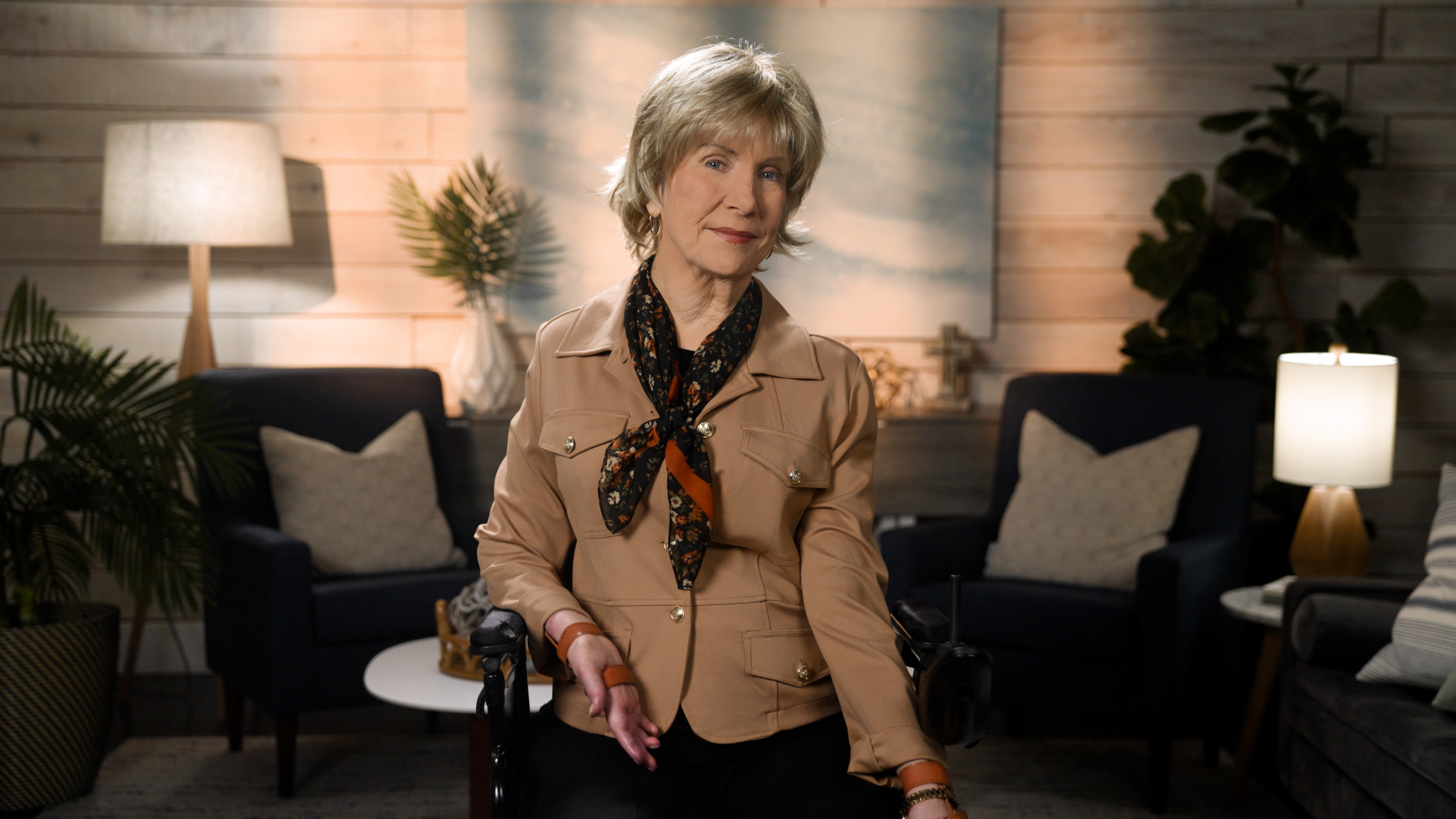 Joni Eareckson Tada smiling for the camera while wearing a brown safari jacket, a black scarf with floral details, in a cozy living room.