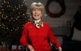 Joni wearing a red sweater with a colorful Christmas-themed pin on her right side and a scarf. She is in a cozy living room with a Christmas tree and gifts in the background, which is slightly blurred.