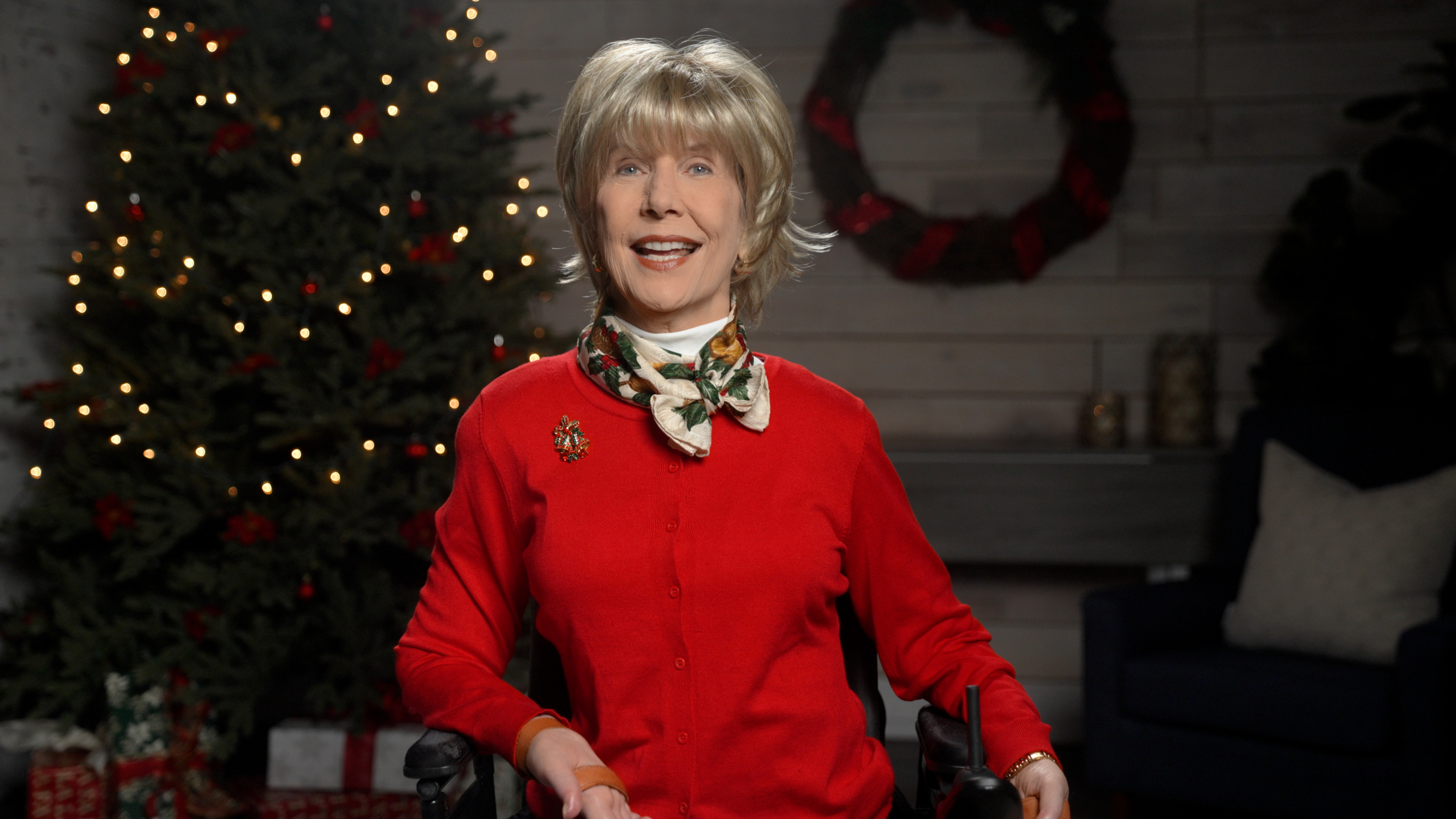 Joni wearing a red sweater with a colorful Christmas-themed pin on her right side and a scarf. She is in a cozy living room with a Christmas tree and gifts in the background, which is slightly blurred.