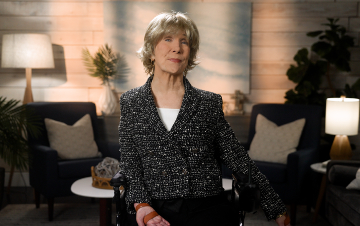 Joni Eareckson Tada smiles for the camera while sitting in her wheelchair, wearing a black blazer with white boucle over a white top, in a cozy living room setting.