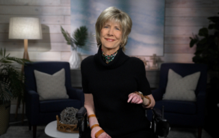 Joni Eareckson Tada smiles for the camera, wearing a black blouse and colorful scarf, with a blurred living room backdrop.