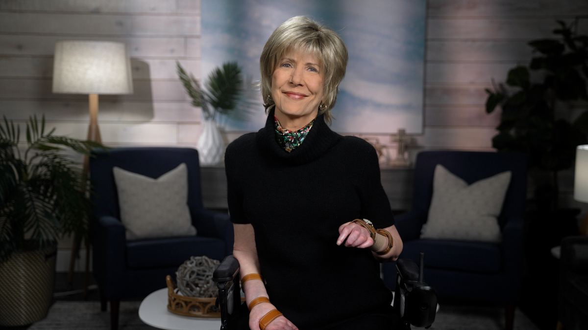 Joni Eareckson Tada smiles for the camera, wearing a black blouse and colorful scarf, with a blurred living room backdrop.