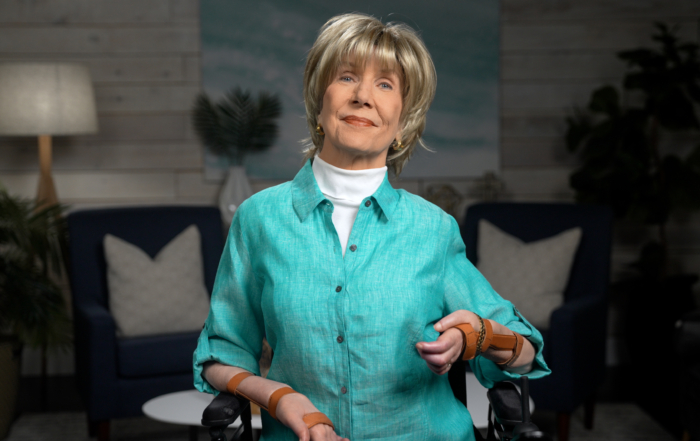 Joni is smiling at the camera, wearing a teal button-up shirt over a white turtleneck, with a cozy, slightly blurred living room in the background.