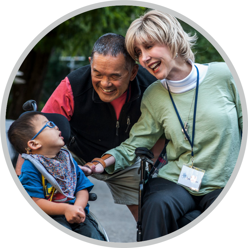 A picture of Joni seated in her wheelchair saying hi to a little boy seated in his wheelchair with Ken beside them leaning down and smiling.