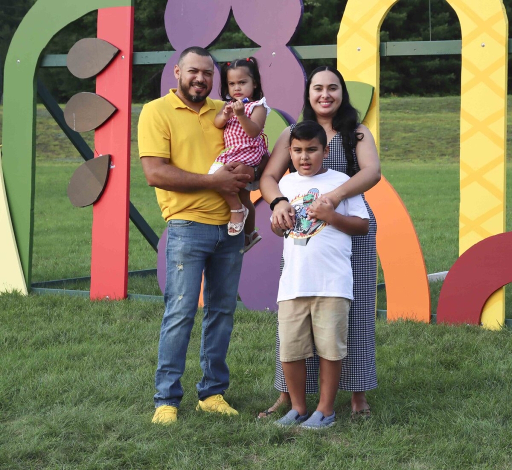 A joyful family photo featuring Oscar, dressed in a yellow shirt and carrying Carin, standing on the right. Luindy, positioned next to Oscar, is gently hugging him. The family is smiling and standing together on lush green grass, with a colorful structure in the background adding vibrancy to the scene.