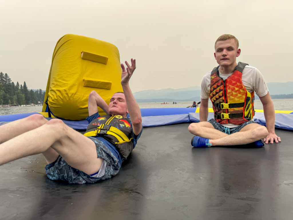 Dino and Tyson enjoying themselves on a trampoline surrounded by water, with a stunning backdrop of pine trees and mountains. They are both wearing life vests, having fun in a beautiful outdoor setting.