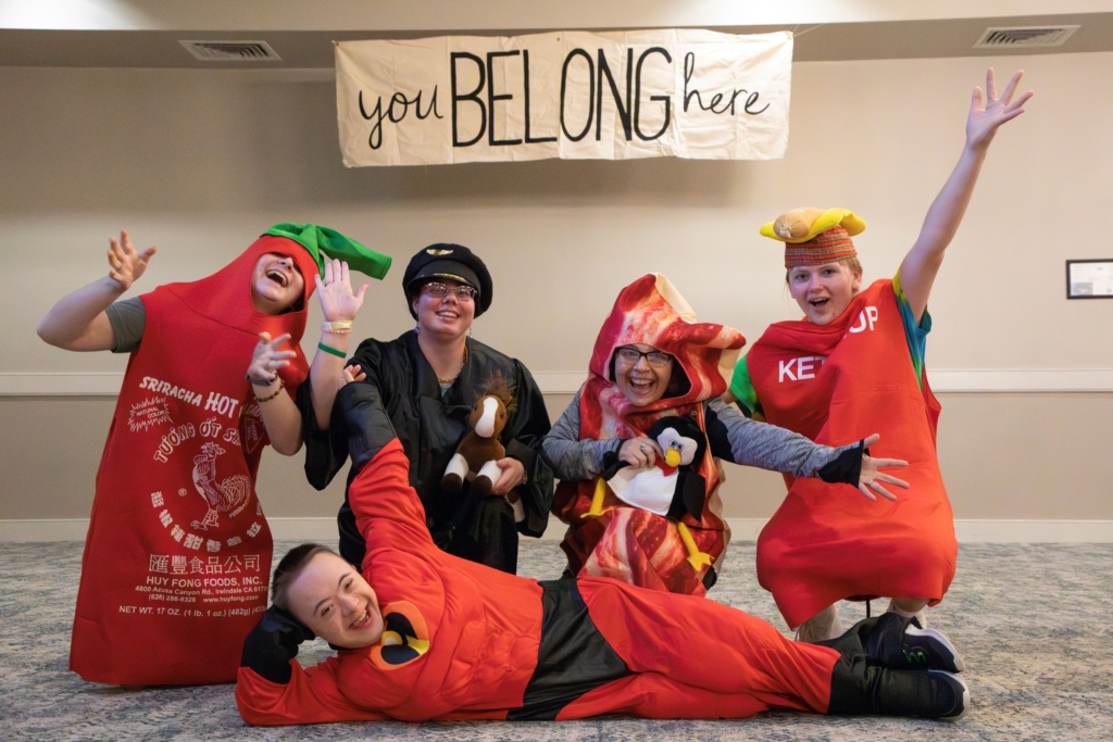 Josiah, Julie, and three other friends wearing costumes. Josiah is dressed in an Incredible costume. A tarpaulin hangs from the ceiling with the text "You Belong Here." Everyone appears happy and is enjoying the moment.