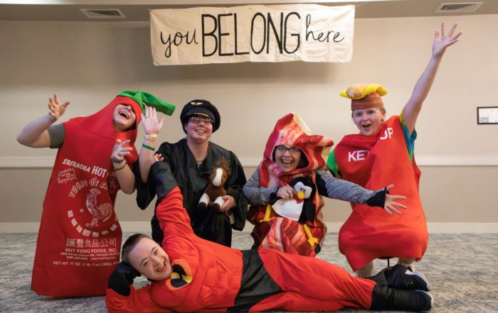 Josiah, Julie, and friends wearing costumes. Josiah is dressed in an Incredible costume. A tarpaulin hangs from the ceiling with the text "You Belong Here." Everyone appears happy and is enjoying the moment.