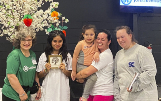 Kimi, a little girl, smiles happily next to Larissa, who is carrying Luiza, with Lilly beside them. All are smiling and posing joyfully for the camera.