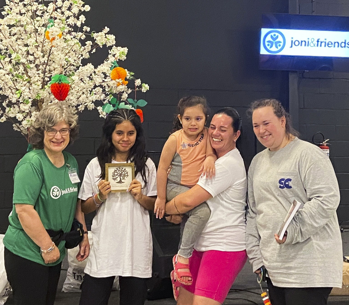 Kimi, a little girl, smiles happily next to Larissa, who is carrying Luiza, with Lilly beside them. All are smiling and posing joyfully for the camera.