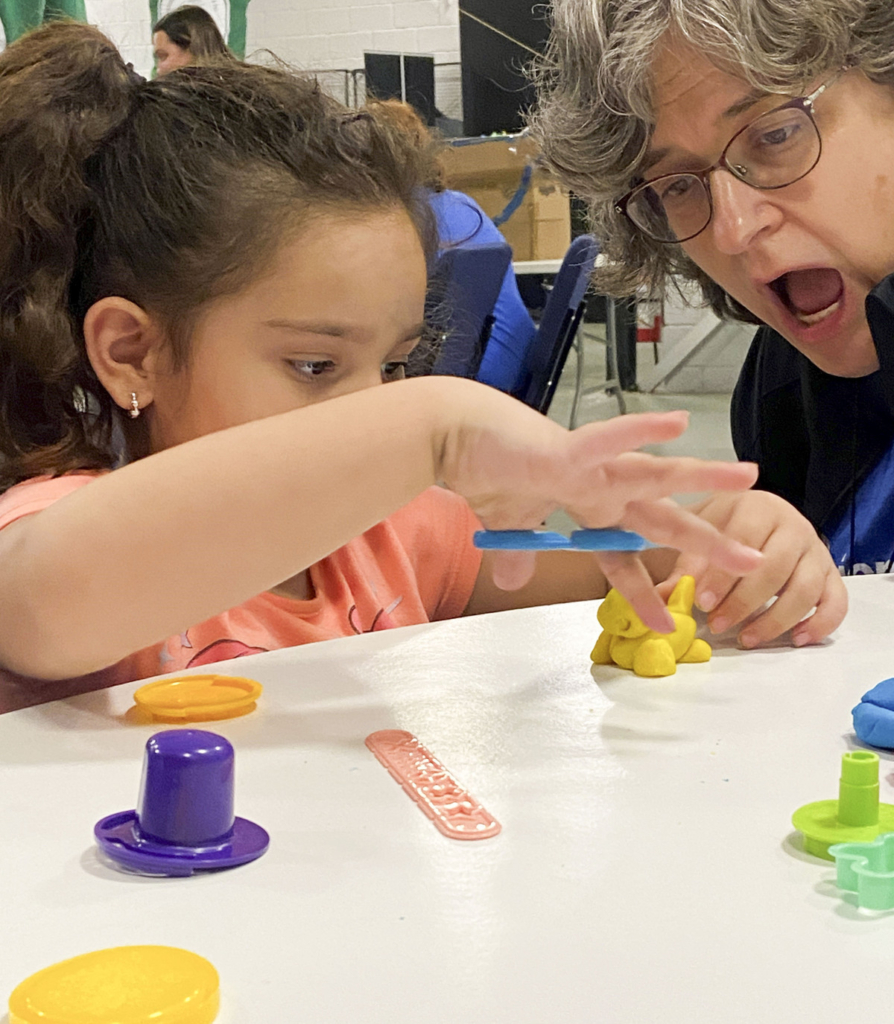 Luiza looks focused as she shapes clay animals, while Kimi, sitting next to her, looks amazed by the clay animal Luiza is making.