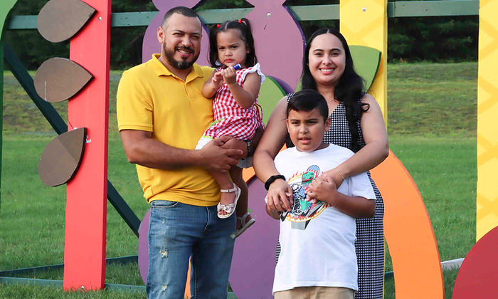 A joyful family photo featuring Oscar, dressed in a yellow shirt and carrying Carin, standing on the right. Luindy, positioned next to Oscar, is gently hugging him. The family is smiling and standing together on lush green grass, with a colorful structure in the background adding vibrancy to the scene.