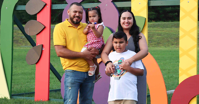 A joyful family photo featuring Oscar, dressed in a yellow shirt and carrying Carin, standing on the right. Luindy, positioned next to Oscar, is gently hugging him. The family is smiling and standing together on lush green grass, with a colorful structure in the background adding vibrancy to the scene.