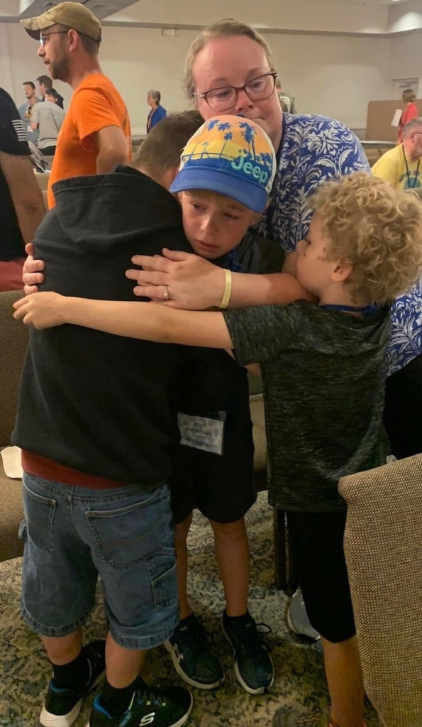 Josiah and Joshua hugging each other, with another child and a woman hugging them as well. They are all sad and crying as they say goodbye.