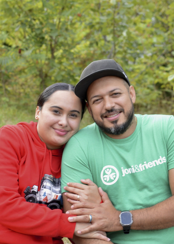 Luindy, wearing a red sweater, is hugging the arm of Oscar, who is wearing a green shirt with a Joni & Friends logo. Oscar is holding Luindy’s hand, and both are wearing noticeable beautiful wedding rings. They are smiling sweetly for the camera, with a backdrop of tall green trees.