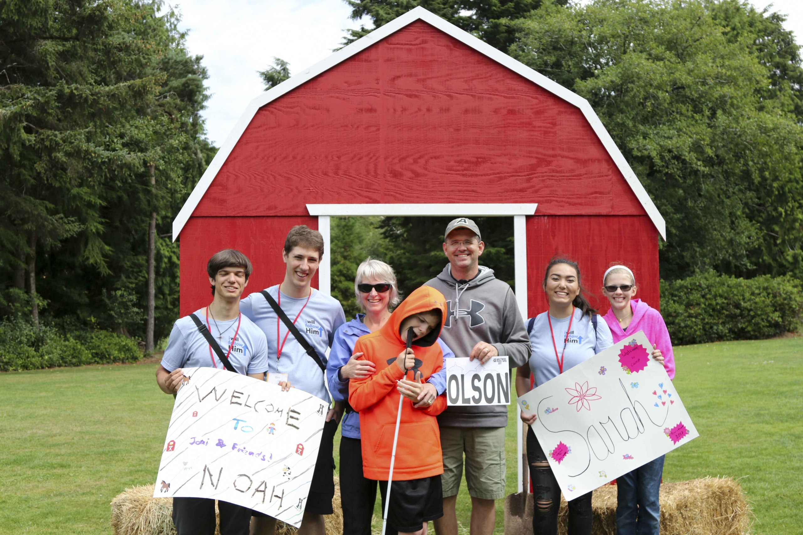 JJ, Will, and Noah smiling for the camera at Family Retreat, surrounded by new friends. Will holds a paper that reads "Joni and Friends Twin Rocks Camp 2017 OLSON." Their friends are holding banners that say, "Welcome to Joni & Friends Noah" and "Welcome to Sarah." Everyone is smiling happily together.